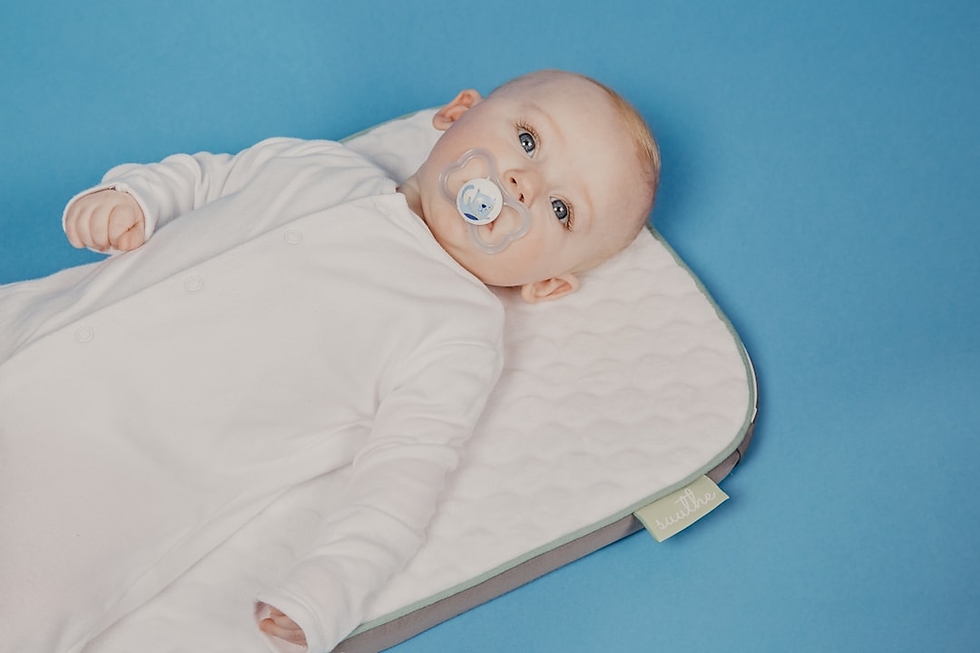 Baby in white onesie with pacifier, lying on a quilted mat. Blue background, calm expression. Tag reads "snuuthe."