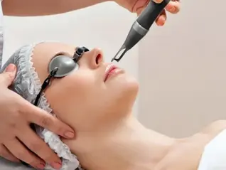 Person lying down with protective goggles, receiving a laser treatment. Technician's hands gently support the head. Clean, clinical setting