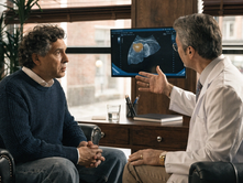 A doctor in a white coat discusses a liver scan on a monitor with a concerned man in a blue sweater. Office setting with a plant and window.