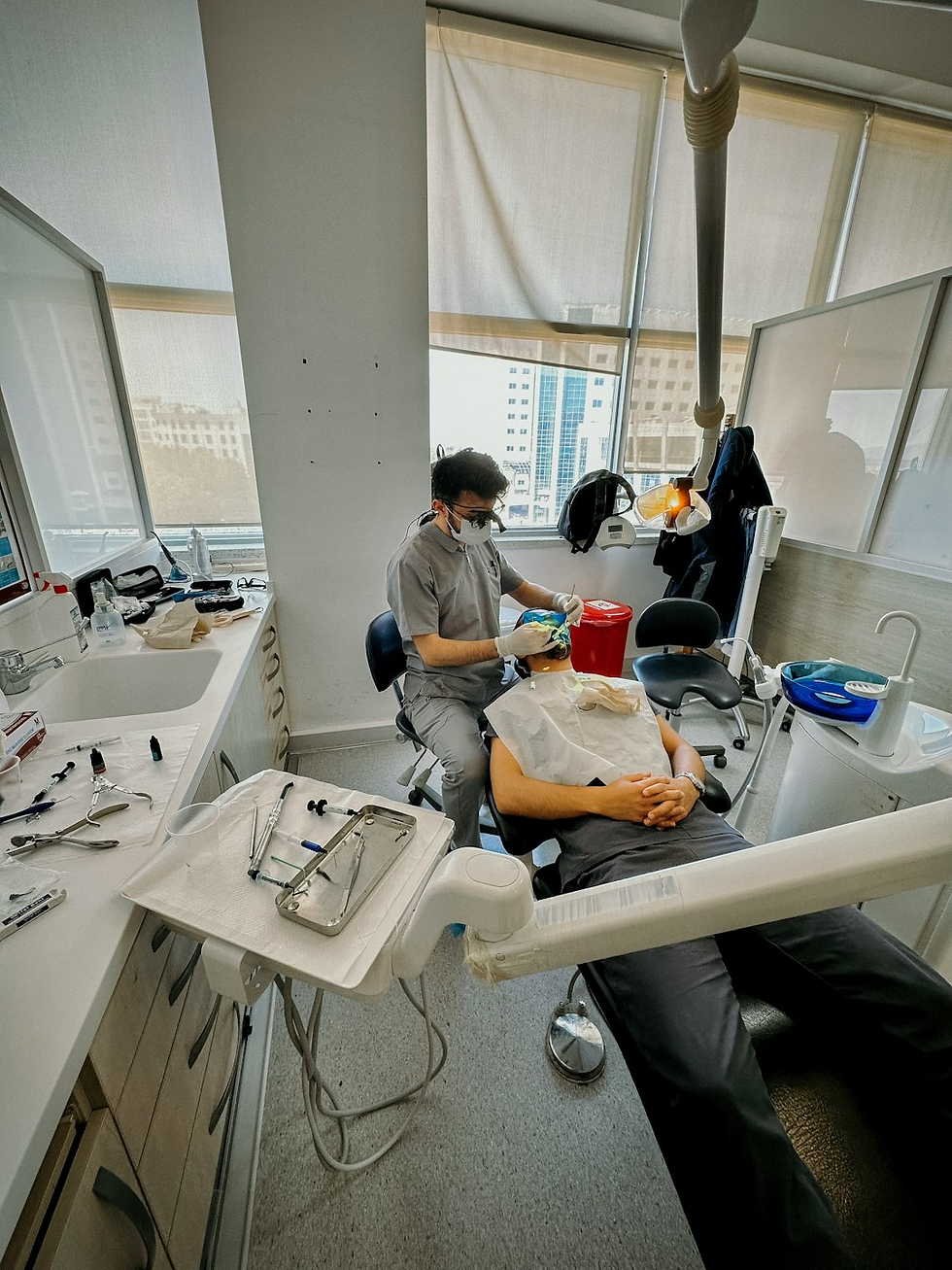 Dentist in gray scrubs working on a patient's teeth in a modern clinic with tools on the counter; bright natural light from large windows.