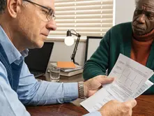 Two people in an office, one holding papers with graphs, the other listening. Books, laptop, and lamp on a wooden table. Serious mood.