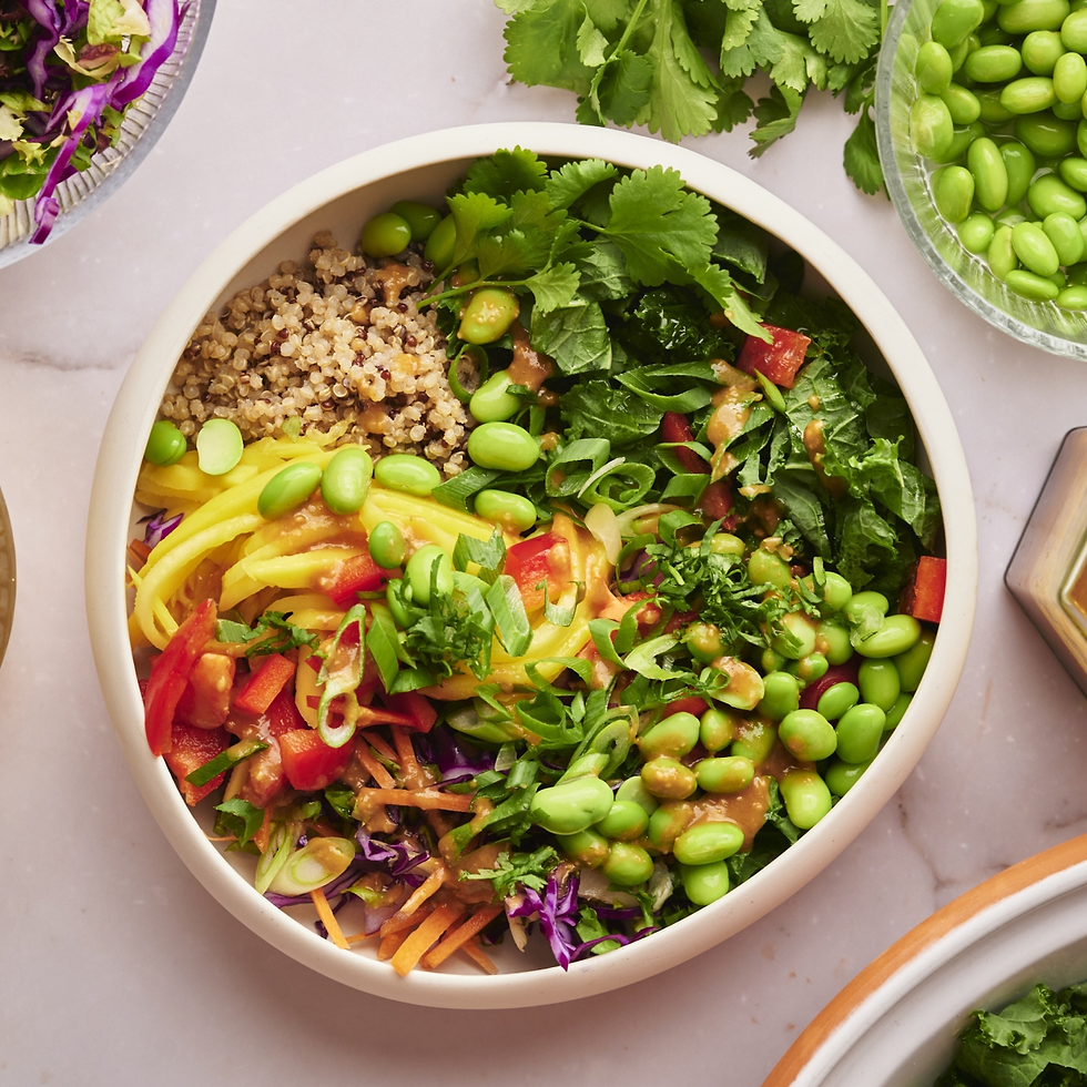 A colorful bowl of quinoa, edamame, sliced peppers, greens, and shredded veggies on a marble surface, surrounded by fresh ingredients.