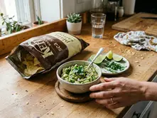 Person serves guacamole in a kitchen with a wooden countertop, Kettle chips bag, lime, herbs, and potted plants near a window.
