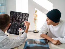 Doctor shows brain scans to a patient in a medical office. A skeleton model and spine sample are on the table. Bright, clinical setting.