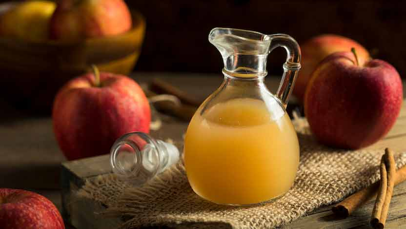 A glass jug of apple cider sits on burlap. Surrounding are red apples and cinnamon sticks on a wooden table, creating a cozy, rustic feel.