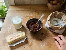 Wooden counter with a bowl of cocoa, glass of milk, butter with "Land O'Lakes," flour bag, whisk, eggs, and plants by window. A hand is visible.