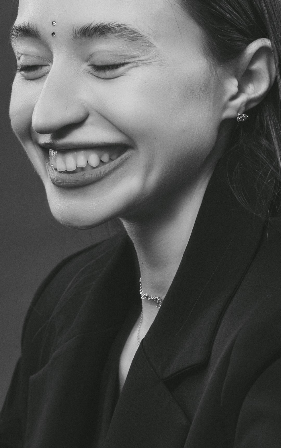 Smiling person with closed eyes, wearing earrings and a necklace, in a black-and-white setting. Joyful mood.