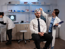 A man sits with EEG cap while a scientist adjusts it. Another person uses a microscope. Lab setting with screens and scientific equipment.