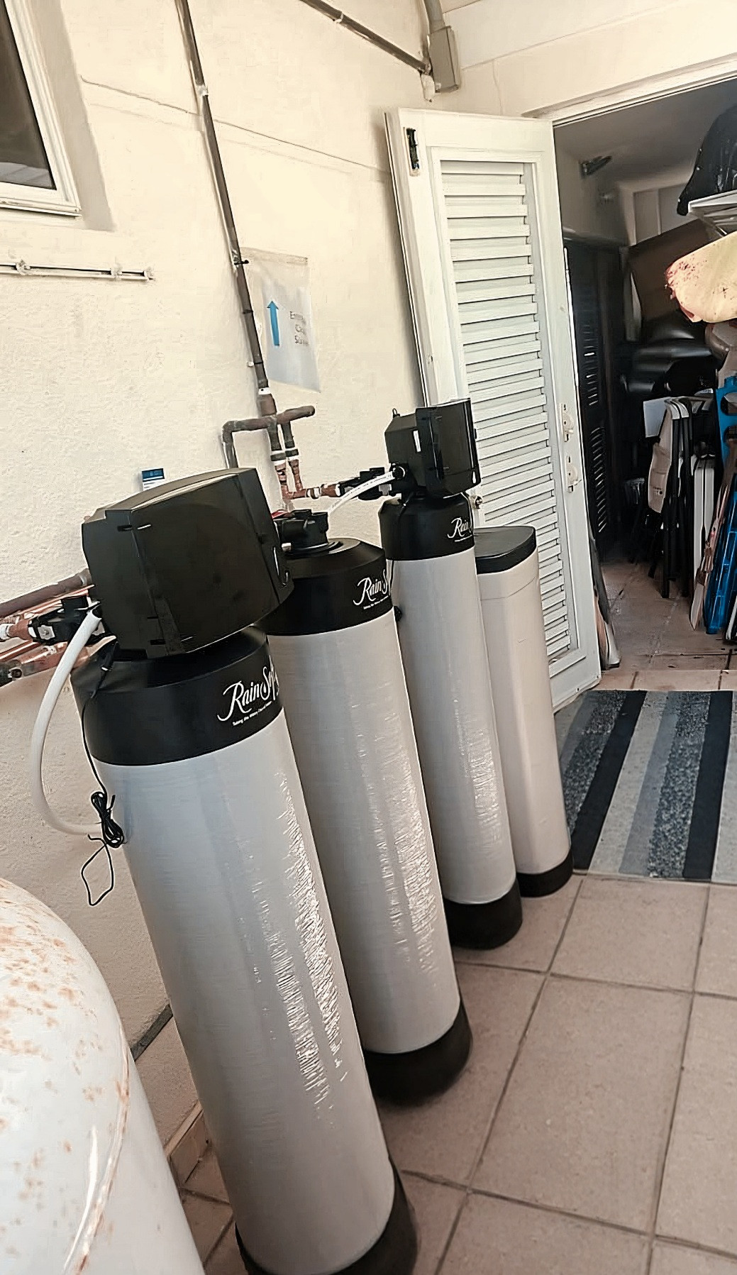 Cuatro tanques de filtración RainSoft en un pasillo de azulejos, junto a una puerta abierta y tuberías en la pared.