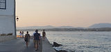Family of five walking seaside road in Spetses, young girl balancing on raised concrete wall, Peloponnese in background, Family Holiday in Greece.