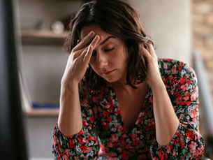 A woman sitting indoors with her eyes closed, holding her head and appearing stressed, overwhelmed and emotionally exhausted.