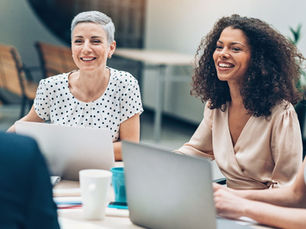 A group of colleagues smiling and chatting around laptops during a collaborative workplace meeting.