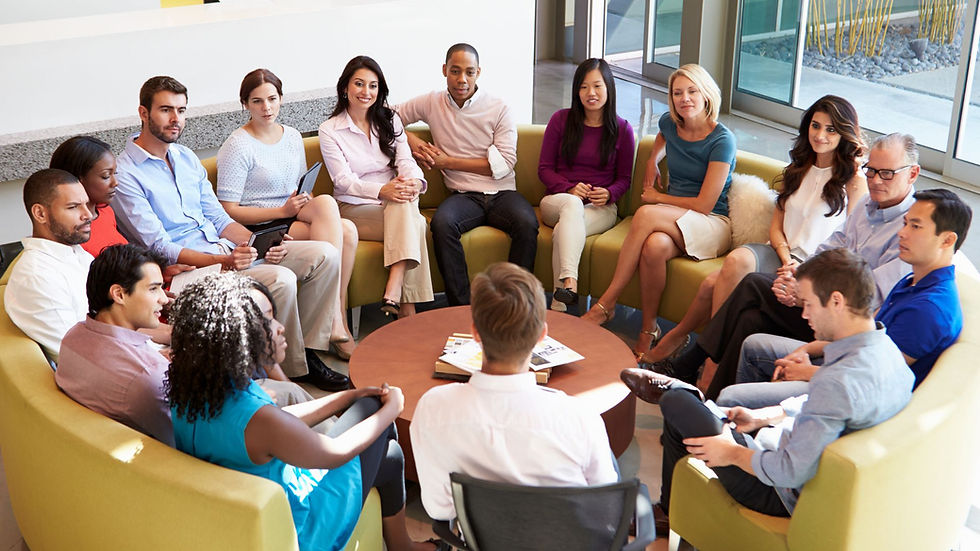 A diverse group of professionals sit in a circle on sofas in a modern office, engaged in a team discussion or meeting. Some hold tablets or notes, while others listen attentively, creating a collaborative and open atmosphere.