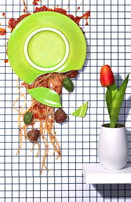 Zerbrochener grüner Teller mit Spaghetti, Fleischbällchen und Tomatensoße, verteilt auf einer karierten Fliesenwand, daneben eine rote Tulpe in weißer Vase. Kreative Foodfotografie von David Weimann aus Köln für Projekte in NRW.

