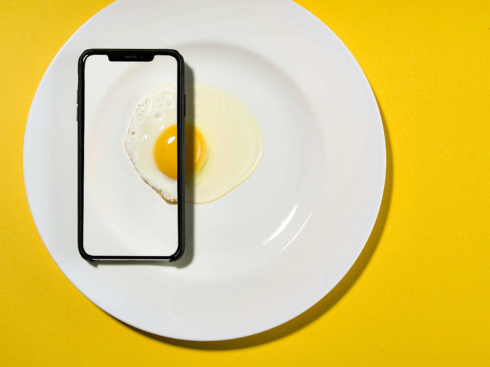 Spiegelei auf weißem Teller mit einem Smartphone darüber, das Eigelb und Eiweiß nahtlos auf seinem Bildschirm zeigt. Kreative Food- und Produktfotografie von David Weimann aus Köln für Markenpräsentationen in NRW.

