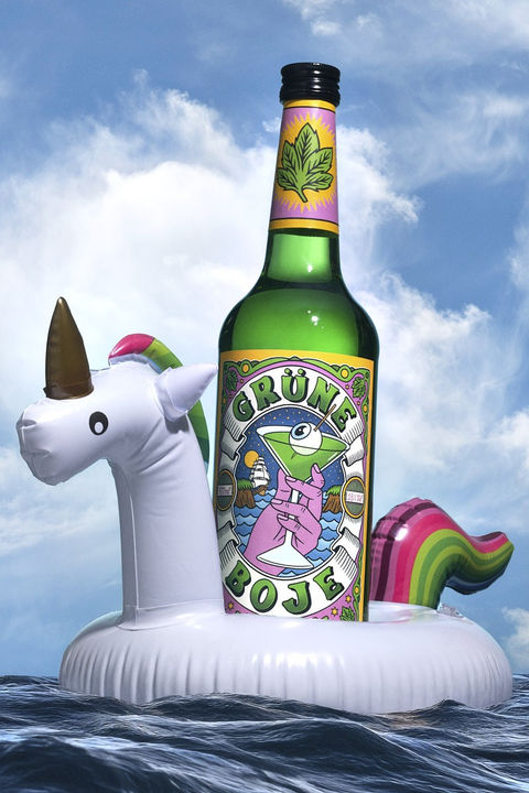 Eine grüne Spirituosenflasche mit bunter Etikettgestaltung auf einem aufblasbaren Einhorn, treibend auf dem Meer unter blauem Himmel. Professionelle Drinkfotografie von David Weimann aus Köln für kreative Markeninszenierungen in NRW.