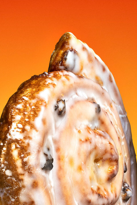 Makroaufnahme einer glasierten Zimtschnecke vor leuchtend orangem Hintergrund.