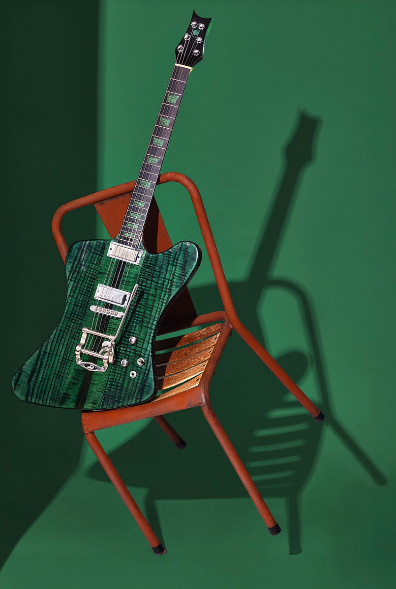 Boutique E-Gitarre mit geflammter Ahorndecke, Bigsby-Tremolo und grüner Lackierung, inszeniert auf Vintage-Stuhl im Studio.