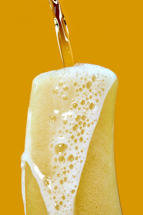 Nahaufnahme eines Bieres, das in ein Glas gegossen wird, mit üppigem Schaum und gelbem Hintergrund. Erfrischende Drinkfotografie von David Weimann aus Köln für kreative Projekte in NRW.