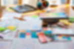Design workspace with sketches, color swatches, laptop, and design tools scattered on a wooden desk.
