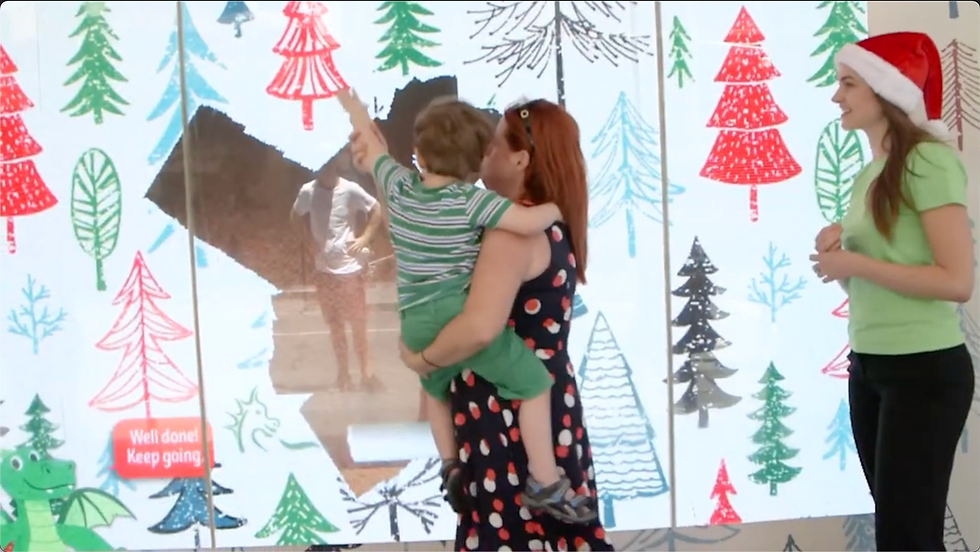 A mother and child interact with an animated Christmas retail window featuring colourful trees and playful graphics, while a staff member in a Santa hat looks on. The interactive display encourages engagement and creates a memorable in-store brand experience.