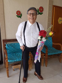 foto de corpo inteiro do personagem, senhor Astolfo, um senhor de 70 anos, com calça social preta, camisa social branca de mangas compridas, sapato marrom e suspensório preto e com uma boina cinza. Na mão direita ele se apoia sobre em uma bengala e na outra mão um pequeno ramalhete de rosas vermelhas e amarelas e folhagens embrulhados em um papel estampado branco e rosa. Ele usa óculos e tem um leve sorriso mostrando os dentes.