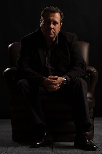 I'm sitting in a dark brown leather armchair, dressed in a black suit, with a lead-colored shirt underneath, no tie, with a watch on my left wrist. I have my hands clasped and my forearms resting on my knees and looking at the camera. The light is dramatic and comes in from the right side.