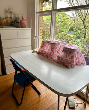 a large white table sits infant of a big window. there are sewing machines under a sheer pink fabric that sit on the table. 