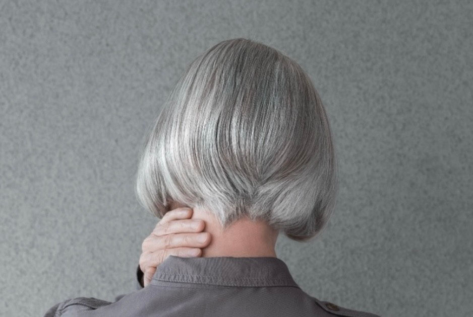 ショートヘアのシルバーヘアの人物が後ろ向きで首を押さえています。背景は灰色の壁で、全体に落ち着いた雰囲気です。