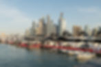 Dubai International Boat Show in Dubai Harbor. Yachts docked at a marina with people walking on a red carpet. Tall buildings rise in the background under a clear blue sky.