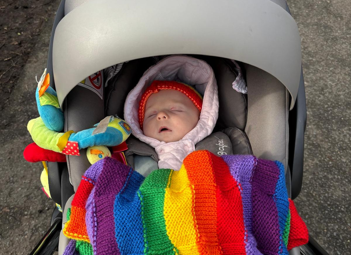 Sleeping baby in a stroller outdoors, tucked under a bright rainbow knit blanket and surrounded by toys.
