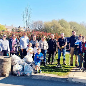 Group of people in a park cleaning up trash; bags filled with litter are piled in front. Sunny day with trees and houses in the background.