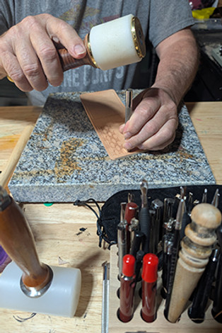 Artisan hand-tooling a piece of leather with a mallet and stamping tool, surrounded by leatherworking tools on a workbench – RLR Leather craftsmanship process
