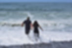 A couple holds hands, walking into the ocean waves on a sandy beach. Both wear black outfits. The sky and sea are blue and expansive.