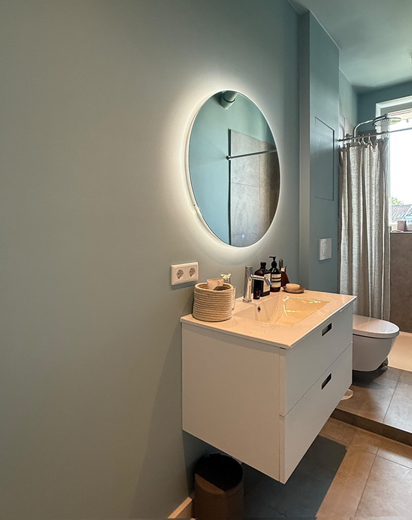 Modernes Badezimmer mit beleuchtetem rundem Spiegel, weißem Waschtisch und blauer Wand. Neutrale Farbpalette, ruhige Atmosphäre.