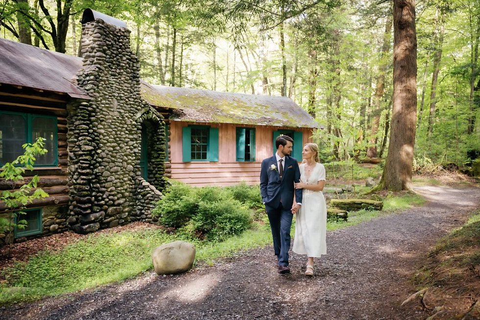 A couple in wedding attire walks on a forest path beside a rustic cabin with a stone chimney. Lush greenery surrounds them, creating a peaceful mood.