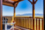 A wooden porch with a chair and drink faces a scenic mountain view under a clear blue sky. Peaceful and relaxing atmosphere.