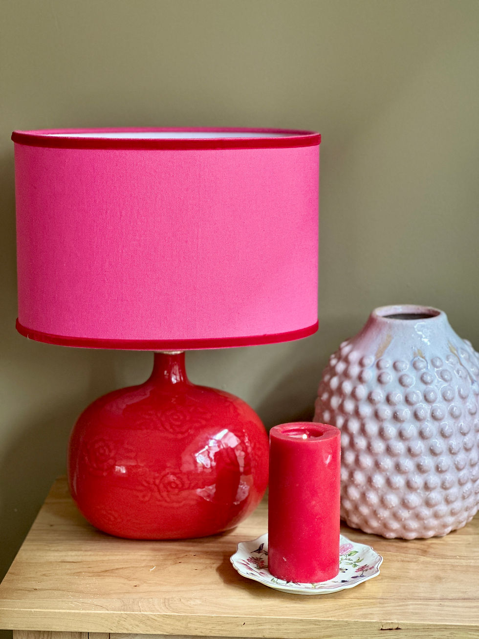Handmade ceramic table lamp with a glossy red base and a soft pink fabric shade, styled on a wooden surface with decorative o