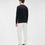 Miniature : Homme portant un pull noir et un pantalon blanc devant un fond clair.