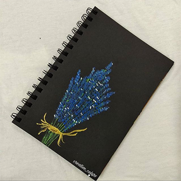 A black notebook lays on a white surface with a drawing of a bundle of purple lavender on the front.