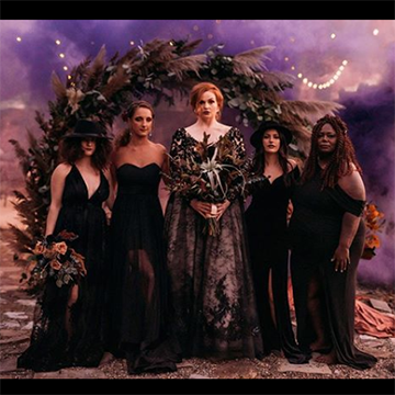 A bride and her bridesmaids stand in black dresses under an arch of flowers and grasses in front of a witchy, purple night sky background.