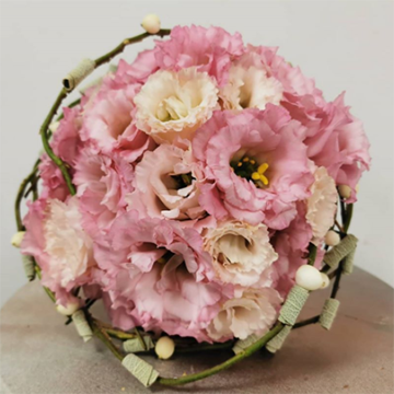 A tight round bouquet of pink and light peach lisianthus flowers sits on a wooden table. We view the bouquet from its top. It has green vines or twigs around its base as decoration.