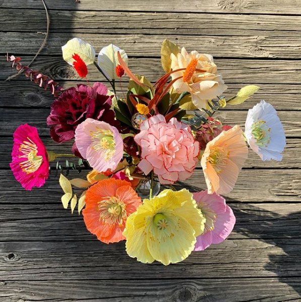 A bouquet of yellow, orange, pink, white, and red poppies with various grasses sits on a background of muted wood texture.