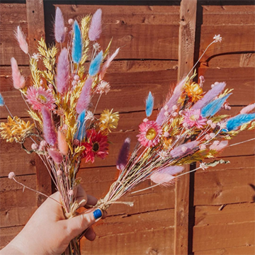 A hand with blue finger nails holds two small sprigs of dried grasses and wheat and small flowers in bright blues, pinks, and purples against a wood background.