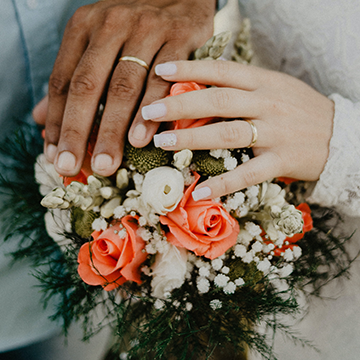 A couple holds their peach and white rose bouquet with baby's breath in between the blooms and the bride holds her hand on top of the bouquet to display her ring. The groom does the same.
