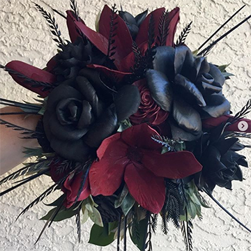 A bouquet of wooden flowers (black roses, burgundy flowers, and black ferns, among others) is held in front of a textured, white wall.