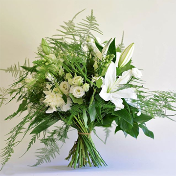 A bouquet stands on its own stems with one large, white lily, a lot of fern greenery, and some smaller white flowers all in front of a white background.