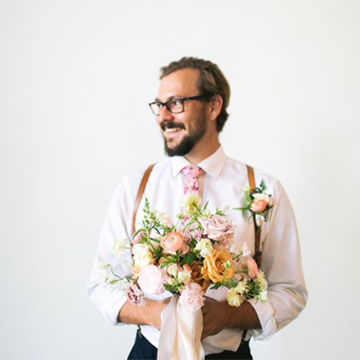 A man with black glasses on turns his head to the side and smiles while wearing a pink tie and suspenders and holding a bouquet with a peach ribbon handle, yellow garden roses, light pink roses, peach and white ranunculus, and various greenery.