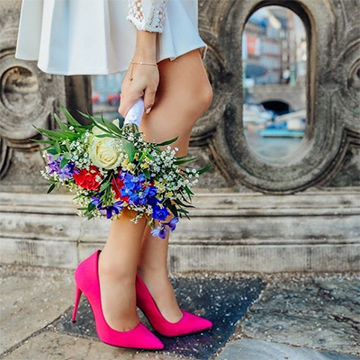 A woman in pink pointed heels holds a colorful bouquet of roses, baby's breath, and various blue and red flowers near her calves while standing in front of an old concrete bridge or structure of some sort.