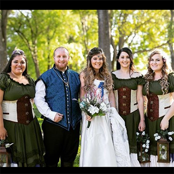 A bride holds her bouquet with her groom (in a blue medieval vest) and her bridesmaids (in green dresses with white and brown corsets, holding hooded lanterns) in the bright woods.